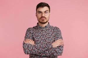 Pensive young bearded male wearing glasses in colorful shirt thinking over something, standing with hands crossed one brow raised questioning, having serious and puzzled expression over pink wall photo