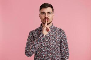 Concerned man wearing glasses with dark hair bearded in colorful shirt, demonstrates a gesture of silence, secret, holding an index finger near the mouth, isolated over pink background photo