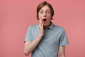 Indoor photo of young man with surprised, amazed expression and dropped jaw isolated on pink background. Guy is shocked by some movie scene, hand touching face