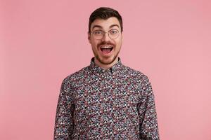 Portrait of a happy excited attractive handsome young man wearing glasses with dark hair unshaved with beard and mustache in colorful shirt opened mouth in amazement, isolated over pink background photo