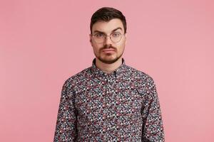 Portrait of suspicious pensive young bearded male wearing glasses in colorful shirt thinking over something, one brow raised questioning, having serious and puzzled expression over pink background photo