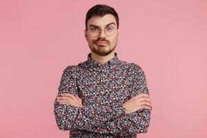 Looks skeptical young bearded male in glasses, colorful shirt thinking over something, standing with hands crossed one brow raised questioning, having perplexed and puzzled expression over pink wall photo