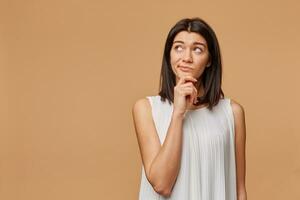 Unsatisfied thoughtful tanned woman standing hand holding chin, frowning, looking at the upper left corner at the blank copyspace, expressing suspiciousness and misunderstanding, over beige background photo