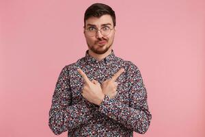 Young man in colorful shirt looking throught glasses having doubts and with confuse face expression keeps hands crossed ond pointing to bouth sides with inde fingers isolated on pink background photo