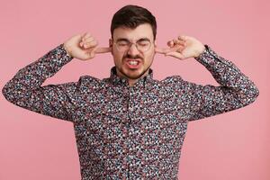 cerca arriba de enojado joven barbado hombre vestido en vistoso camisa, mantiene cerrado ojos, dos dedos cerca su orejas, demostración gesto de sordera, ignora alguien, terminado rosado pared foto