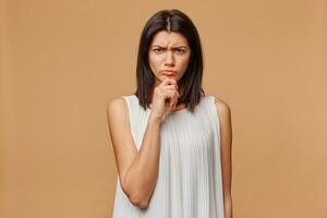 Indoor photo of serious suspicious tanned woman standing hand keeps chin, frowning, expressing distrustfulness and concern, over beige background