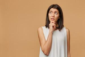 Thoughtful tanned woman standing hand holding chin looking at the upper left corner at the blank copyspace with a little caution, portrays the excitement, over beige background photo