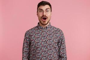 Amazed attractive handsome young man wearing glasses with dark hair unshaved with beard and mustache in colorful shirt opened mouth in excitement, isolated over pink background photo