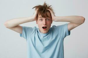 Portrait of shocked amazed young man with braces on teeth, hands on head and opened mouth wears blue t shirt feels astonished and stunned isolated over white background photo