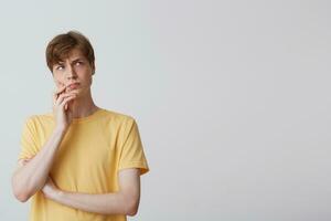 Closeup of thoughtful handsome young man wears yellow t shirt standing, keeps hands folded and thinking isolated over white background photo