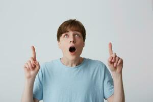 Portrait of surprised astonished young man with opened mouth wears blue t shirt feels surprised and points up to copyspace with both hands isolated over white background photo