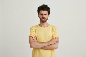 Portrait of serious strict bearded young man wears yellow t shirt feels angry and keeps arms crossed isolated over white background photo