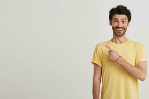 Closeup of cheerful handsome bearded young man wears yellow t shirt smiling and points to the side with finger isolated over white background photo