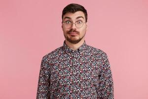 Surprised man stares through spectacles with misunderstanding, bewilderment, wearing colorful shirt shrug shoulders in uncertainty, over pink background photo