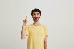 Portrait of smiling attractive bearded young man wears yellow t shirt looks happy and points up by finger isolated over white background photo