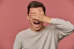 Close up of young shocked guy in blank long sleeve, stands over pink background with wide open eyes and screaming, covers eyes with palm. photo