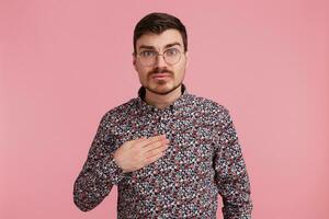 Uncertain surprised man in spectacles wearing colorful shirt pointin with palm on himself tries to make situation clear, over pink background photo