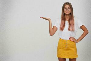 Closeup of amazed happy young woman wears t shirt, yellow skirt and spectacles looks surprised and holds copyspace on palm isolated over white background photo
