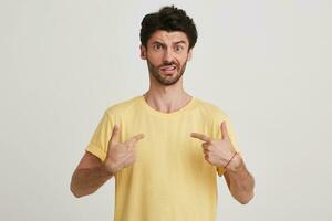Portrait of amazed shocked bearded young man wears yellow t shirt feels astonished and points at himself with both hands isolated over white background photo