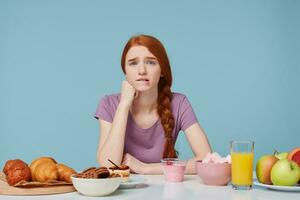 triste Pelirrojo niña mirando cámara picaduras labio, preocupaciones dudas acerca de nutrición, salud, piensa acerca de dieta, extra calorías, horneando comida y Fresco Fruta jugo yogur laico en el mesa, en un azul antecedentes foto