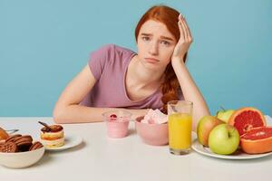 joven Pelirrojo niña se sienta a el mesa con su cabeza en su mano tristemente entiende el importancia de Fresco Fruta y sano alimento, pero quiere a comer pastel, aislado en un azul antecedentes foto