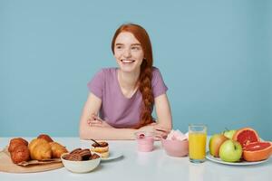 Studio shot of smiling red-haired girl with braided hair sitting at a table, about to eat lunch looking leftside. On the white table lay baking and fresh healthy food. photo