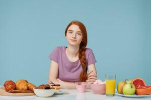 A beautiful red-haired girl with braided hair sitting at a table, about to eat breakfast looking at the camera. On the table baking on one side and on the other fresh fruit juice yogurt.Healthy food photo