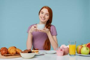 Red-haired girl sitting at a table with slightly bowed head, smiles holds white cup with delicious drink in hands, has lunch looking camera. On the table baking products and fresh food lay photo