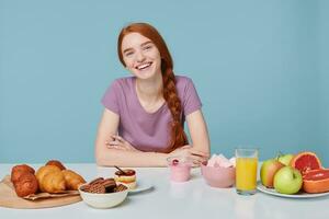 Young attractive red-haired girl smiles dreamily looks to the upper right corner, sitting at a white table on which are fresh, healthy products and delicious pastries baking products. photo