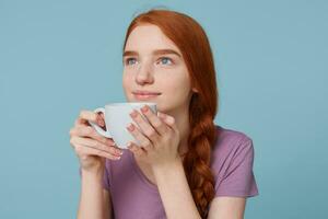 Close up of lovely beautiful red-haired girl smiling dreamingly looking upper left corner,keeps in hands big white cup with drink, over blue background photo