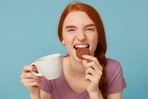 cerca arriba de un encantador hermosa Pelirrojo niña sonriente en broma mirando cámara, mantiene en mano blanco taza con beber, con apetito picaduras chocolate Galleta, terminado azul antecedentes foto