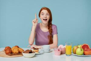 joven linda inspirado emocionado Pelirrojo niña sentado a el mesa durante desayuno. un bueno idea ocurrió a su, elevado su índice dedo hacia arriba, en el mesa laico pasteles y Fresco sano productos foto