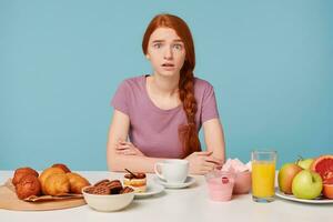 A red-haired girl is frightened, sitting at the table with arms folded, in front lay baking croissants and healthy foods and fruits, do not know what to choose. photo