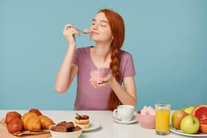 linda Pelirrojo niña molesto saboreo Cereza yogur, cerrado su ojos desde placer, lame un cucharilla sentado a el mesa durante almuerzo, pasteles en el mesa y Fresco fruta, en contra un azul antecedentes. foto