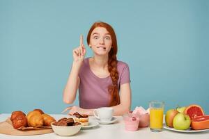 A lovely girl is sitting at a table during lunch time, a good idea suddenly came to mind,raised index finger. On the table is pastries and fresh, healthy products, fruits,isolated on a blue background photo