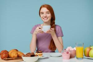 sonriente Pelirrojo niña con trenzado pelo sentado a un mesa, sostiene blanco taza con delicioso bebida en manos, tiene almuerzo mirando cámara. en el mesa horneando productos y Fresco comida laico foto