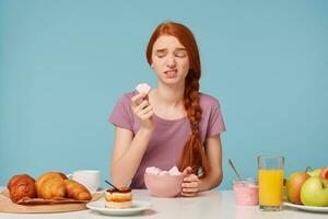 niña sentado a un mesa durante desayuno intentó un Fruta malvavisco, participación un pedazo en su manos mira a él con asco, ella no me gusta el gusto aislado en un azul antecedentes. foto