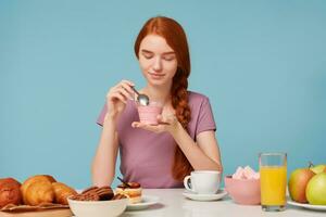 A cute red-haired girl with a braid sits at a table, has lunch, holds in her hands a cherry yogurt, looks at him within temptation and is going to eat with a teaspoon. photo