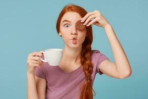 cerca arriba de un gracioso hermosa Pelirrojo niña en broma mirando cámara, mantiene en mano blanco taza con beber, cubre ojo con chocolate Galleta, hace pescado cara con labios, terminado azul antecedentes foto