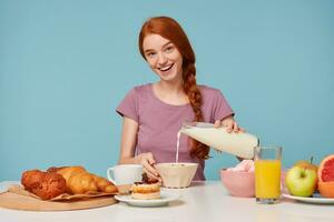 Cheerful kind cheerful red-haired girl sitting at the table is going to have breakfast. Pour milk into a plate with mussels. Smiling affably, looking at the camera. Isolated on a blue background. photo