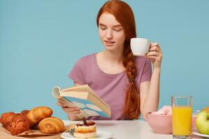 Close up of charming red-haired girl with braided hair, sitting at a table, drinks from white cup delicious tea, has breakfast reading book. On table baking products and fresh food lay,over blue wall photo
