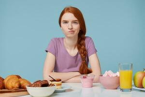 Photo of red-haired girl looking camera with anger discontent, doubts thinks about diet, extra calories, baking food and fresh fruit juice yogurt lay on the  table, isolated on a blue background