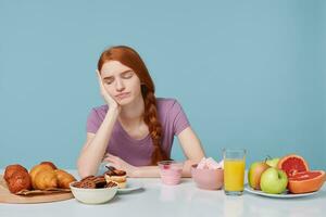 estudio Disparo de Pelirrojo niña mirando con descontento tristeza en horneando productos piensa acerca de qué comida a comer, Fresco Fruta jugo yogur en el otro lado de mesa, aislado en un azul antecedentes foto