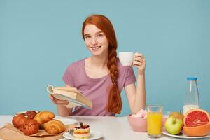 estudio Disparo de atractivo sonriente Pelirrojo niña con trenzado pelo,sentado a un mesa, sostiene blanco taza con delicioso bebida en mano, tiene almuerzo leyendo libro.en mesa horneando productos y Fresco comida laico foto