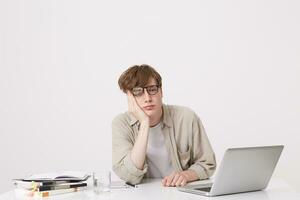 Portrait of sad bored young man student wears beige shirt and spectacles looks tired sitting at the table with laptop computer and notebooks isolated over white background photo