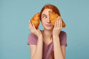 Playful cheerful red-haired girl holding two croissants in her hands, looks away covers closes her eye with one croissant, isolated on a blue background. photo