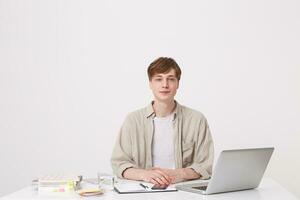 Closeup of confident young man student wears beige shirt sitting and working with laptop computer and notebooks at the table isolated over white background photo