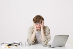 Closeup of sad unhappy young man student wears glasses and beige shirt looks depressed sitting at the table with laptop computer and notebooks isolated over white background photo