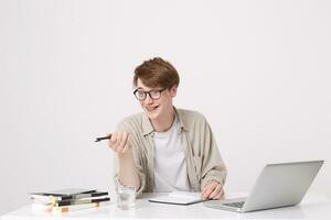 Portrait of cheerful attrative young man student wears glasses and beige shirt points to the side and study at the table with laptop computer and notebooks isolated over white background photo