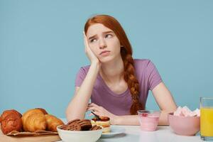 interior foto de triste Pelirrojo niña pensando acerca de alimento, salud, dieta, extra calorías, horneando productos y Fresco Fruta jugo yogur son en el mesa, aislado en un azul antecedentes